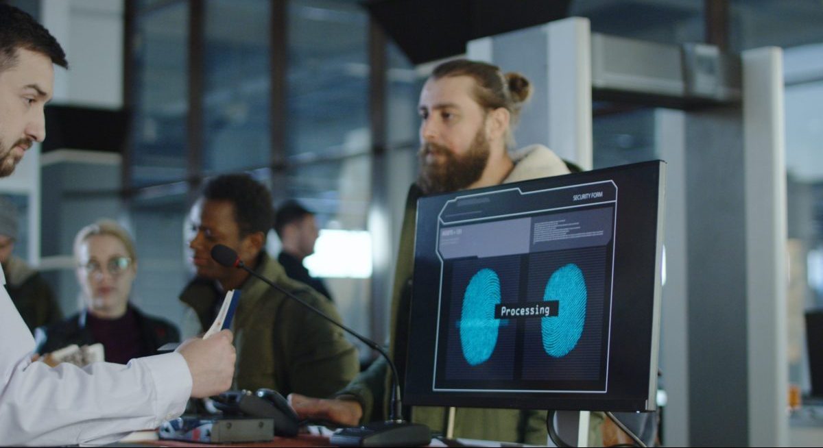 Man giving fingerprints with sensor when passing special control point in airport with employee watching at screen.